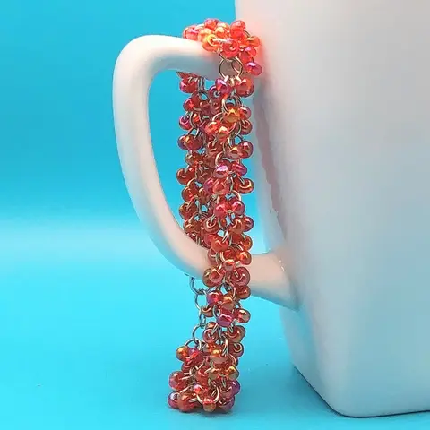 Silver chain mail bracelet with red rainbow beads hanging on a white mug with a blue background