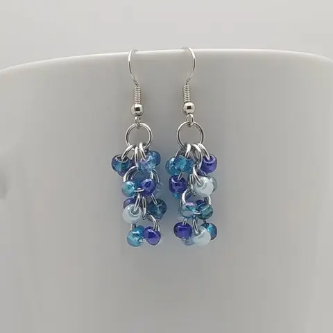 Silver dangly earrings with blue mix beads hanging on a white mug.