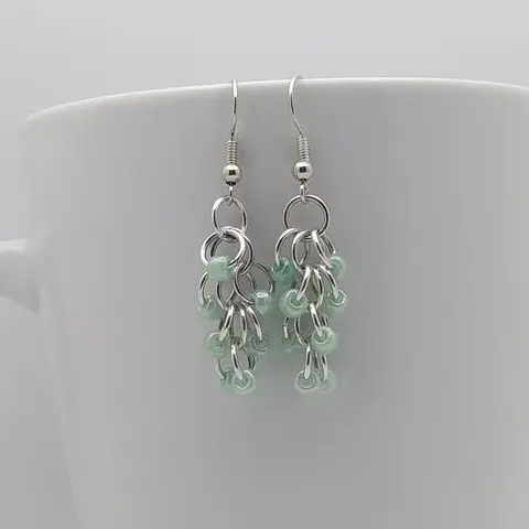 Silver dangly earrings with green beads hanging on a white mug.