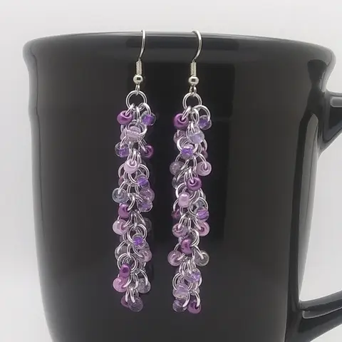 Silver dangly chain mail earrings with purple mix beads hanging on a black mug.