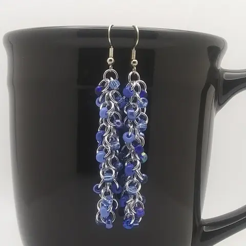 Silver dangly chain mail earrings with sapphire blue mix beads hanging on a black mug.