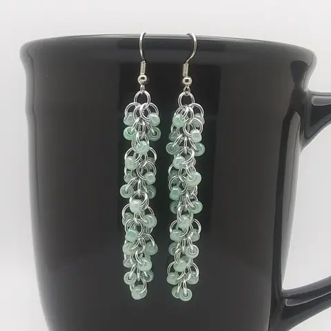 Silver dangly chain mail earrings with green beads hanging on a black mug.