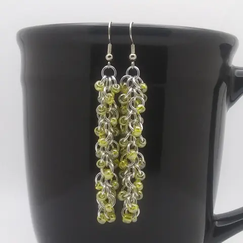 Silver dangly chain mail earrings with yellow beads hanging on a black mug.