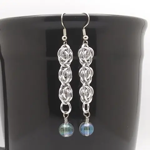 Silver dangly chain mail earrings with green ab glass beads hanging on a black mug.