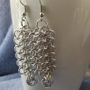 Silver dangly chain mail earrings hanging on a white mug.