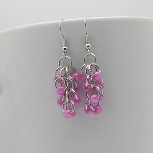 Silver dangly chain mail earrings with dark pink beads hanging on a white mug.