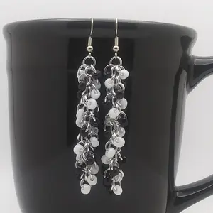 Silver dangly chain mail earrings with a swirl of black and white beads hanging on a black mug.