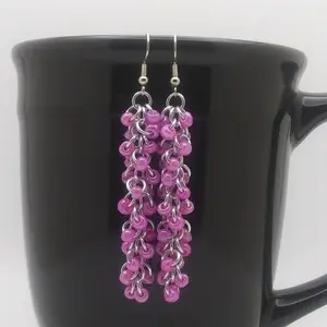 Silver dangly chain mail earrings with dark pink beads hanging on a black mug.