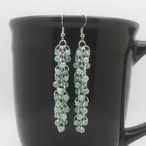 Silver dangly chain mail earrings with green beads hanging on a black mug.