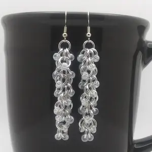 Silver dangly chain mail earrings with crystal luster beads hanging on a black mug.