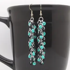 Silver dangly chain mail earrings with a swirl of black and turquoise beads hanging on a black mug.